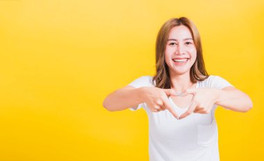 Asya Tayland mutlu portresi güzel güzel genç kadın gülümsemesi parmak kalp figürü şekil işareti yap iki elle ve kamerayla işaret et, stüdyo görüntüsü izole edilmiş sarı arka plan fotokopi alanı ile