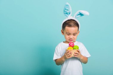 Asyalı şirin çocuk gülümsüyor tavşan kulağı ve beyaz tişört giyiyor. Mavi arka planda paskalya yumurtası dolu bir sepeti tutmak için bekliyor.