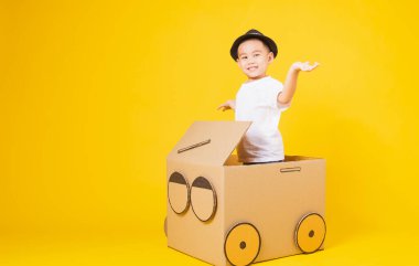 Portre mutlu Asyalı şirin çocuk gülümsüyor beyaz tişört giyip karton araba sürüyor, sarı arka planda fotokopi çekiliyor.