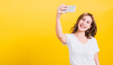 Asya Tayland portresi gülümseyen güzel güzel bir kadın beyaz tişört giyer, selfie fotoğrafı çeker, telefonuna bakan bir video görüşmesi yapar, stüdyo çekimlerinde izole edilmiş sarı arka plan fotokopi çekilir.