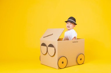 Portre mutlu Asyalı şirin çocuk gülümsüyor beyaz tişört giyip karton araba sürüyor, sarı arka planda fotokopi çekiliyor.