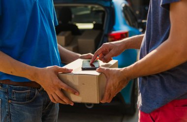 Asyalı genç kurye üniformalı paket posta kutusuyla çevrimiçi alışveriş yapıyor. Koruyucu yüz maskesi takıyor ve evdeki dijital cep telefonunda el parmak izini kullanarak müşteri ve servis yapıyor.
