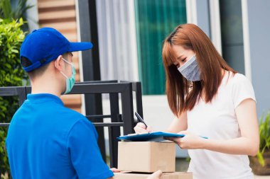 Paket postası taşıyan Asyalı genç teslimatçı, sokağa çıkma yasağı altındaki karantina altındaki kağıt defter üzerindeki kalem imzasını kullanarak koruyucu yüz maskeli kadın müşteri COVID-19 'u karantina altına aldı.