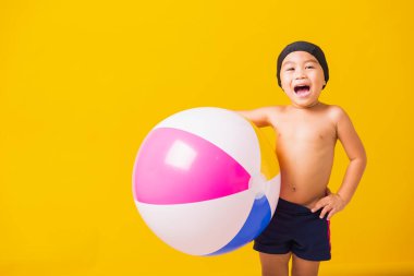 Yaz tatili konsepti, Asyalı mutlu çocuk portresi, mayo içinde gülümseyen çocuk plaj topu, yaz tatilinde şişme topla eğlenen çocuk, izole edilmiş sarı arka plan.