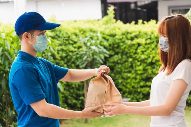 Asyalı kurye genç adam, sokağa çıkma yasağı altındaki karantina altında kadın müşteriye hem koruyucu yüz maskesi hem de karantina altındaki Coronavirus COVID-19 kullanan kadın müşteriye kağıt poşetler veriyor.