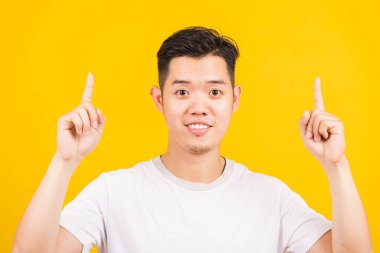 Mutlu Asyalı yakışıklı adam gülümsüyor beyaz tişört giymiş yukarı doğru işaret ediyor kameraya bakıyor, stüdyo görüntüsü izole edilmiş sarı arka plan.