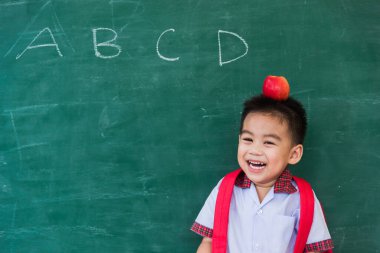 Okula geri dön. Anaokulundan, okul çantasıyla, kafasında kırmızı elmayla yeşil okul tahtasına gülümseyen küçük, mutlu Asyalı çocuk.
