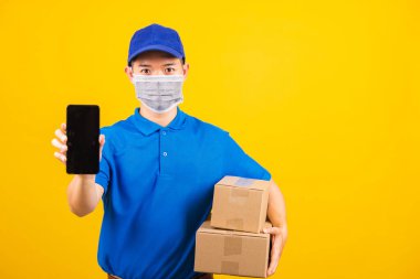 Asya 'lı genç teslimatçı erkek tişörtü ve şapka üniforması giyiyor. Yüz maskesi takıyor. Coronavirus ya da COVID-19 altında siyah akıllı cep telefonu ekranını gösteriyor.