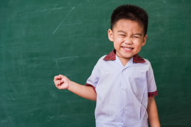 Okula geri dön. Anaokulundan, öğrenci üniforması içinde, yeşil okul tahtasına parmak ucuyla gülümseyen mutlu Asyalı komik, sevimli çocuk.