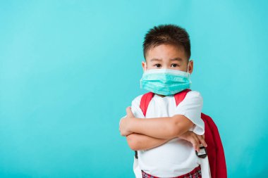Okula geri dön Coronavirus Covid-19 eğitimi. Asyalı küçük çocuk yuvası portresi yüz maskesi takın ve okul çantası okula gitmeden önce çapraz kollar, stüdyo çekimi izole edilmiş mavi arka plan