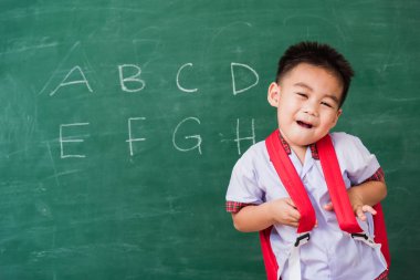 Okula geri dön. Mutlu Asyalı komik küçük çocuk anaokulu okul üniforması içinde okul çantası giyerek yeşil okul tahtasına gülümseyen ilk okul eğitim konsepti.