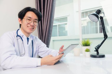 Mutlu Asyalı genç doktor yakışıklı adam gülümsüyor ev ofisinden çalışma gözlükleri takıyor, hastane ofisindeki siyah modern dijital tablet bilgisayar kullanıyor, Teknoloji sağlık ve tıp konseptini kullanıyor.