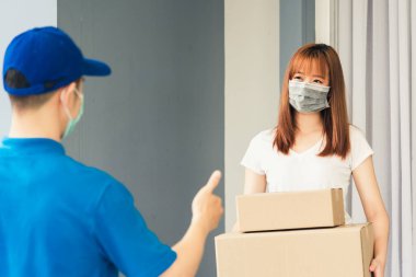 Asyalı kurye genç adam kadın müşteriye paket kutuları veriyor. Hem koruyucu yüz maskesi alıyor hem de sokağa çıkma yasağı pandemik koronavirüs altında iyi bir destek işareti için parmaklarını gösteriyor.
