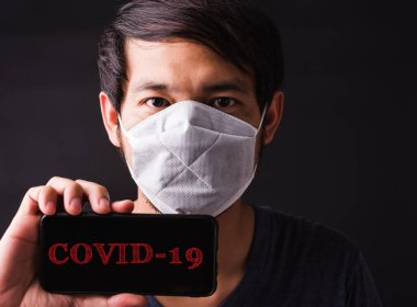Gözlerinde koruyucu yüz maskesi olan Asyalı yakışıklı adam yüzünün yanında cep telefonu boş ekranı, hijyen önleme COVID-19 veya koronavirüs koruma konsepti, siyah arka plan gösteriyor.