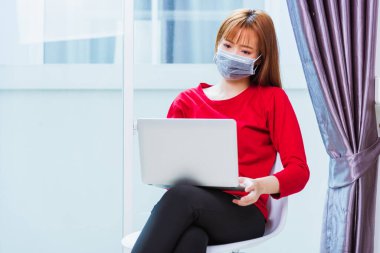 Asyalı genç iş kadını kırmızı tişört giyer yüz maskesi takar ev bilgisayarından çalışır Coronavirus hastalığını karantinaya alır veya COVID-19