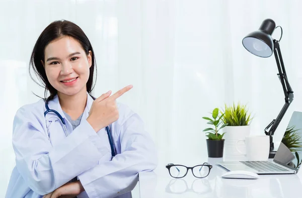 ポートレート病院のオフィスでパーソナルノートパソコンを扱うアジアの自信に満ちた女性医師 タイの女性医療専門家スタッフ笑顔彼女のポイント指でサイドアウェイスペースオンデスクスペースフロントコンピュータ — ストック写真