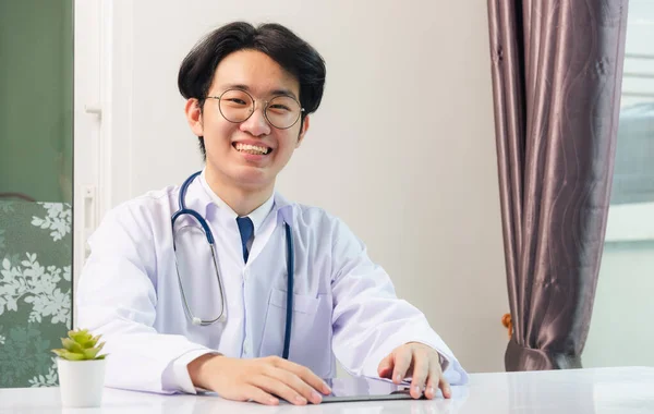 幸せなアジアの若い医師ハンサムな男性の笑顔は 自宅のオフィスからの仕事は 病院のオフィス テクノロジーヘルスケアと医学の概念でデスク上の黒い現代的なスマートデジタルタブレットコンピュータを使用して眼鏡を着用 — ストック写真