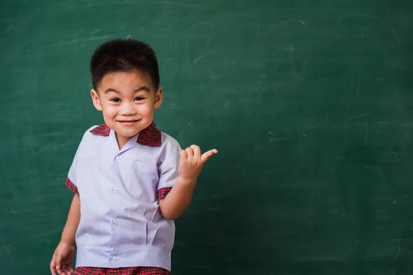 Back to School. Happy Asian funny cute little child boy from kindergarten in student uniform smiling point finger to side away space on green school blackboard, First time to school education concept