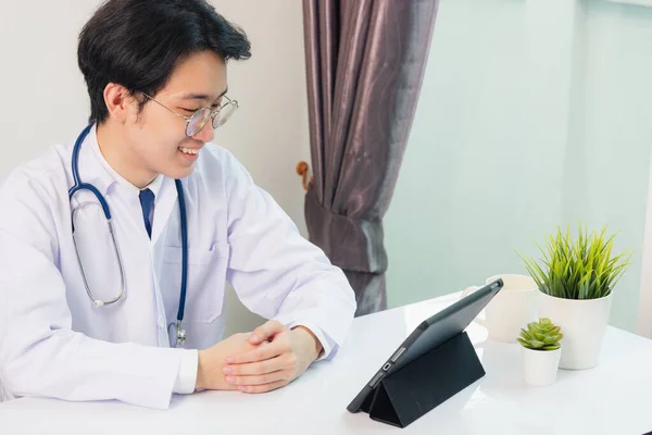 幸せなアジアの若い医師ハンサムな男の笑顔眼鏡を使用して現代的なスマートデジタルタブレットコンピュータ座って 患者が病院のオフィスで机の上に症状を説明聞きます テクノロジーヘルスケアと医学 — ストック写真