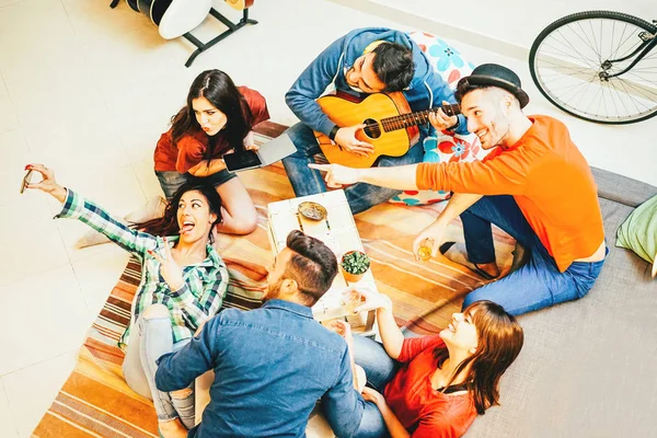 Group of funny friends enjoying together playing music with guitar and taking selfie with mobile phone - Happy young people having fun in the living room at home - Concept of friendship