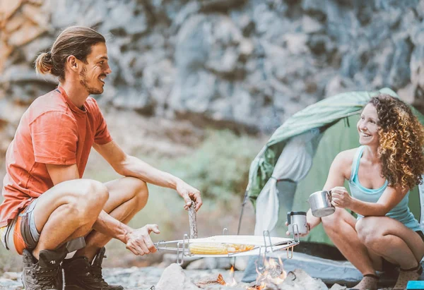 Trekker couple camping in rock mountains with a tent - Climber people cooking and drinking hot tea next to bonfire - Travel, extreme sport, wild life concept