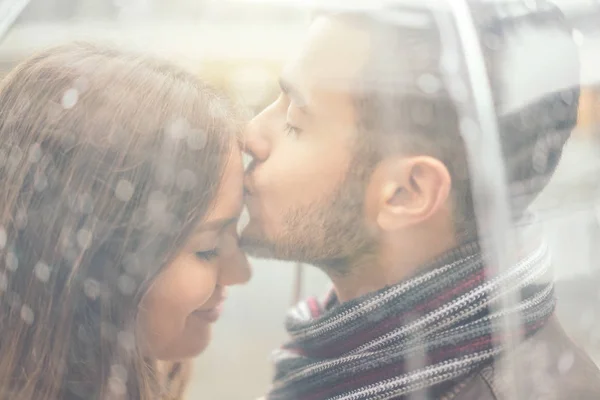 Beautiful young couple having a romantic tender moment under the rain - Handsome man kissing his girlfriend forehead under a transparent umbrella in a rainy day - Concept of Love and relationship