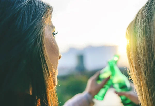 Young women toasting beer on a sunset - Happy friends drinking and cheering beers in a park outdoor admiring the beautiful view - People, drinks, friendship concept - Focus on female face on the left