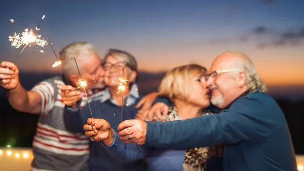 Happy senior friends celebrating with sparklers outdoor - Older people having a fun and tender moment on rooftop - Love, party, elderly lifestyle concept