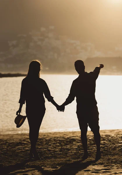 Back view of a beautiful young couple of teenagers holding their hands standing on the beach during sunset - Silhoutte of two lovers having a romantic moment on the beach - Love and vacation concept