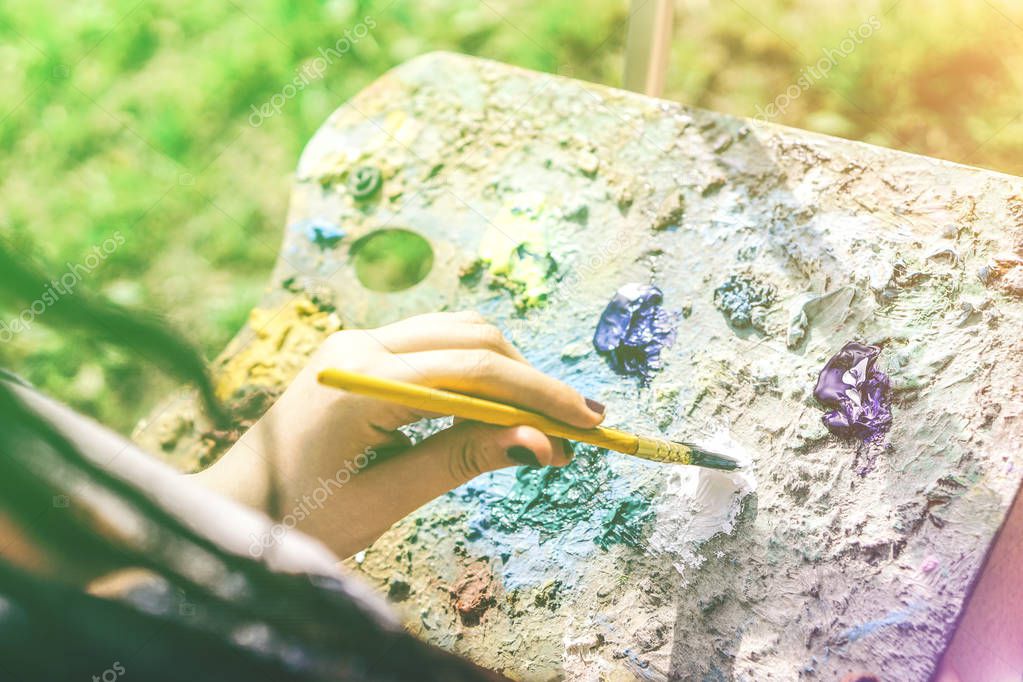 Young artist painting in a park outdoor - Close up of painter with dreadlocks hairstyle working on her art in a colorful garden - Concept of people expressing arts - Focus on hand paint
