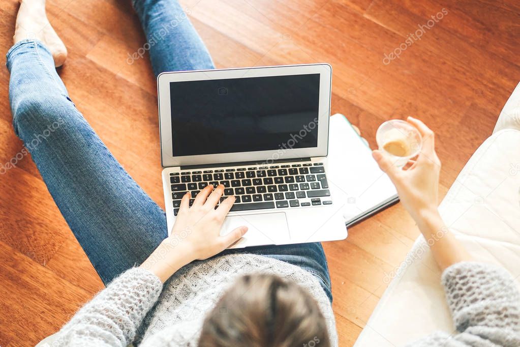 Top view of a young student using her computer while sitting on the floor under sofa holding coffee in her hand - Woman studying on her laptop while drinking coffee in living room - Focus on notebook