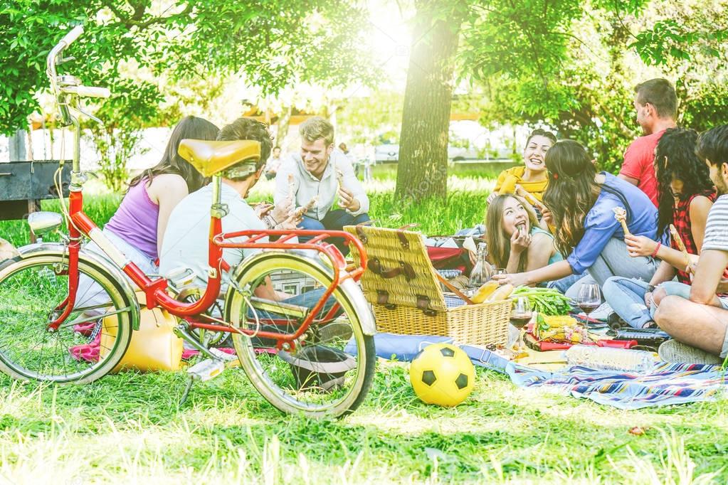 Group of a lot of people having a tasty picnic eating and drinking red wine in a park outdoor - Happy friends celebrating a sunny day sharing drinks and foods - Traditional meal merged with nature