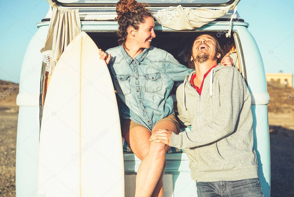 Happy couple of surfers standing behind on vintage camper van - Young people adventuring on road trip with a minivan transport - Concept of travel, hippie, lifestyle