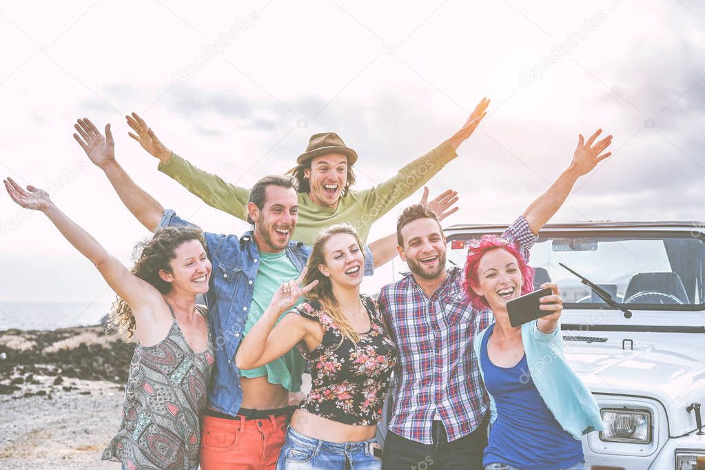 Group of happy friends taking a selfie using mobile smart phone during a road trip with jeep car - Travel people having fun spreading hands up while taking self photos in desert - wanderlust concept