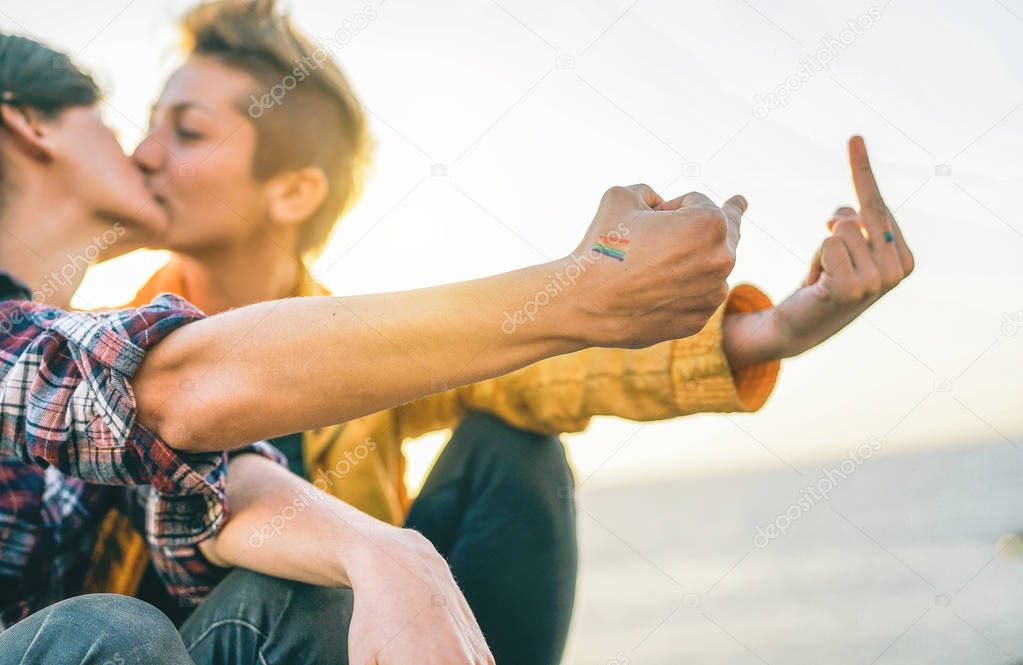 Lesbian couple kissing at sunset - Gay having a tender moment on the beach while making middle fingers up on their hands lgbt flag - Diversity, homosexuality, happiness concept