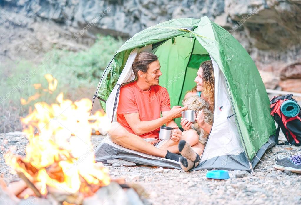 Happy couple of hikers with their dog camping with tent around rock mountains next to fire - People relaxing in a camp drinking hot tea after a trekking day - Travel, love, sport and lifestyle concept