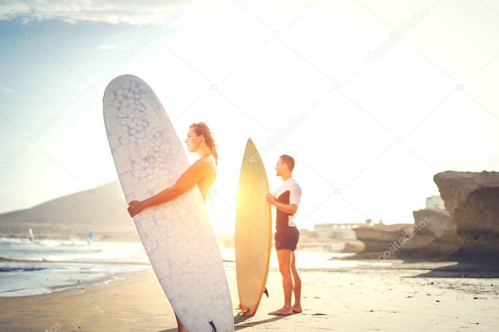 Young couple of surfers standing on the beach with surfboards preparing to surf on high waves during a magnificent sunset behind the mountain - People, lifestyle, sport concept - Focus on woman