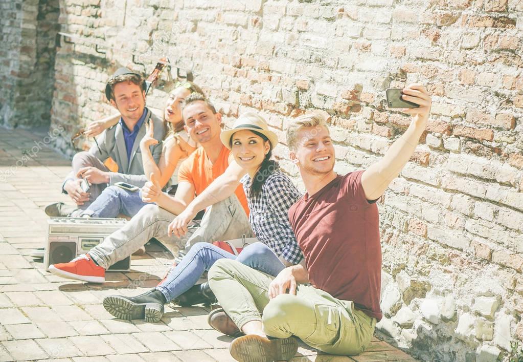 Group of multiracial friends taking a selfie with a mobile smartphone camera - Self portrait of happy persons sitting on the street and listening music with a vintage stereo - Focus on first man