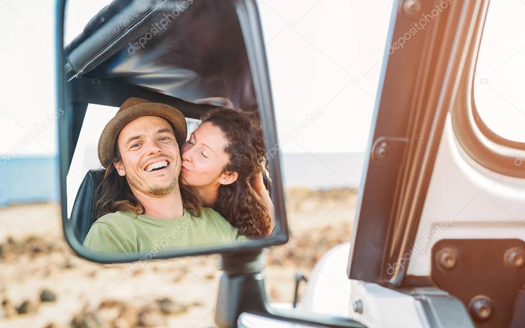 Happy couple having a tender moment during their road trip with a convertible car - Pov in a car mirror of young travel people in their vacation - Love, relationship, traveler lifestyle concept