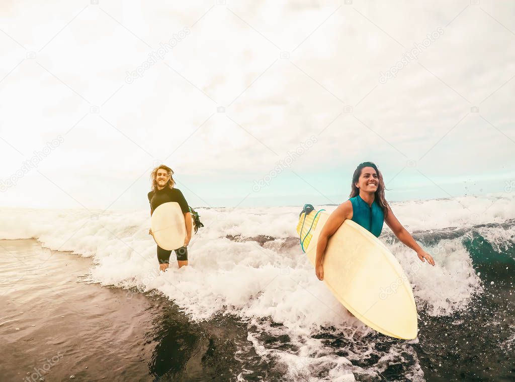 Surfers couple running together with surfboards on ocean at sunset time - Sporty fit friends having fun surfing on high waves - Travel and extreme sport culture lifestyle concept