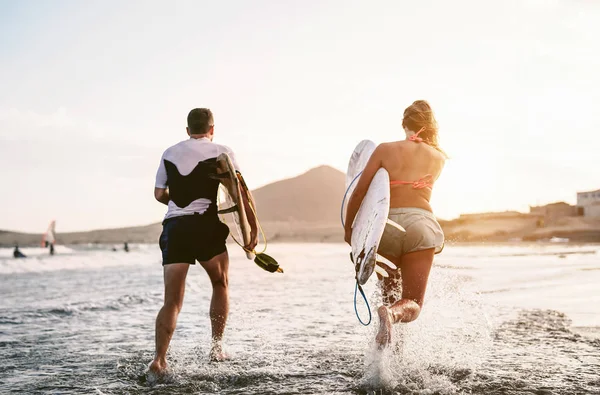 Happy surfers running in the water at sunset time - Young couple having fun surfing in ocean - Extreme sport and youth culture lifestyle concept — Stock Photo, Image