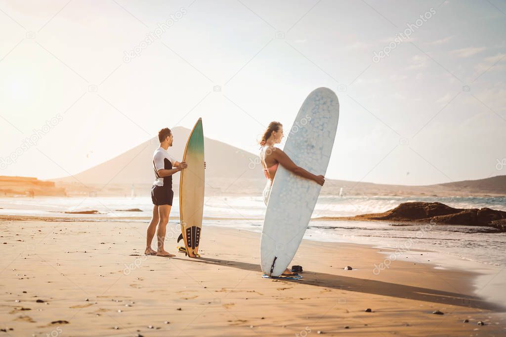 Surfers couple standing on the beach with surfboards preparing to surf on high waves - Young people having fun during surfing day - Extreme health sport and youth lifestyle culture concept