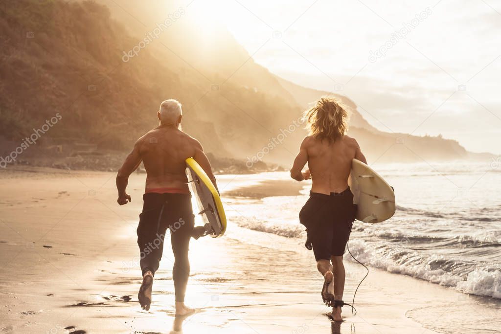 Happy fit friends having fun surfing on sunset time - Surfers father and son running out the ocean - Sporty health people lifestyle and extreme sport concept