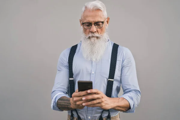 Старший человек, использующий мобильный смартфон и слушающий музыку с воздушными капсулами - Fashion bearded male working on digital marketing social media - Elderly lifestyle people and technology concept — стоковое фото