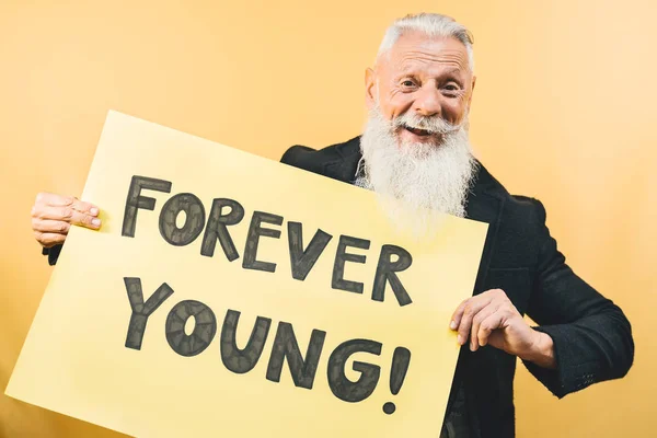 Happy youthful senior feeling forever young - Hipster mature man giving message with yellow banner - Elderly people lifestyle and positive emotions concept — Stock Photo, Image
