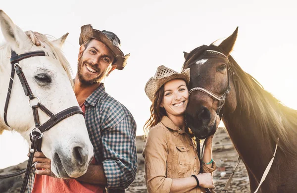 Happy couple having fun with horses inside stable - Young farmers sharing time with animals in corral ranch - Human and animals relationship lifestyle concept