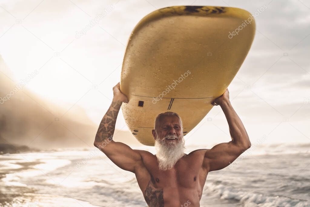 Happy fit senior having fun surfing at sunset time - Sporty bearded man training with surfboard on the beach - Elderly healthy people lifestyle and extreme sport concep