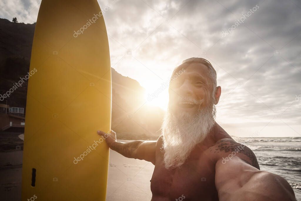 Happy fit senior having fun surfing at sunset time - Sporty bearded man taking selfie while training with surfboard on the beach - Elderly healthy people lifestyle and extreme sport concept