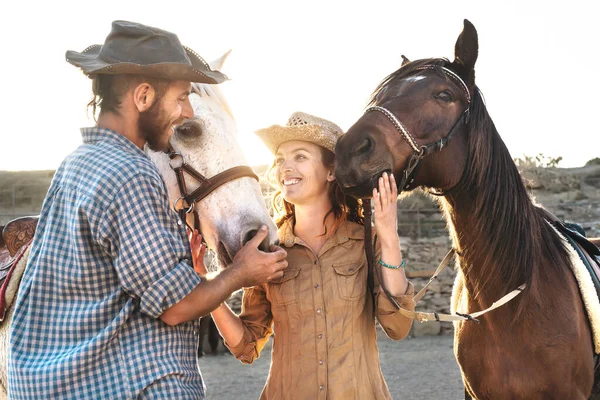 Happy couple having fun with horses inside stable - Young farmers sharing time with animals in corral ranch - Human and animals relationship lifestyle concept
