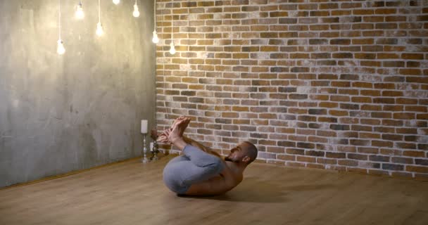 A muscular, lithe, bearded man does yoga in a gym against a brick wall, he sits on a transverse twine and bends his back forward. — Stock Footage A muscular, lithe, bearded man does yoga in a gym against a brick wall, he sits on a transverse twine and bends his back forward. Stock Footage